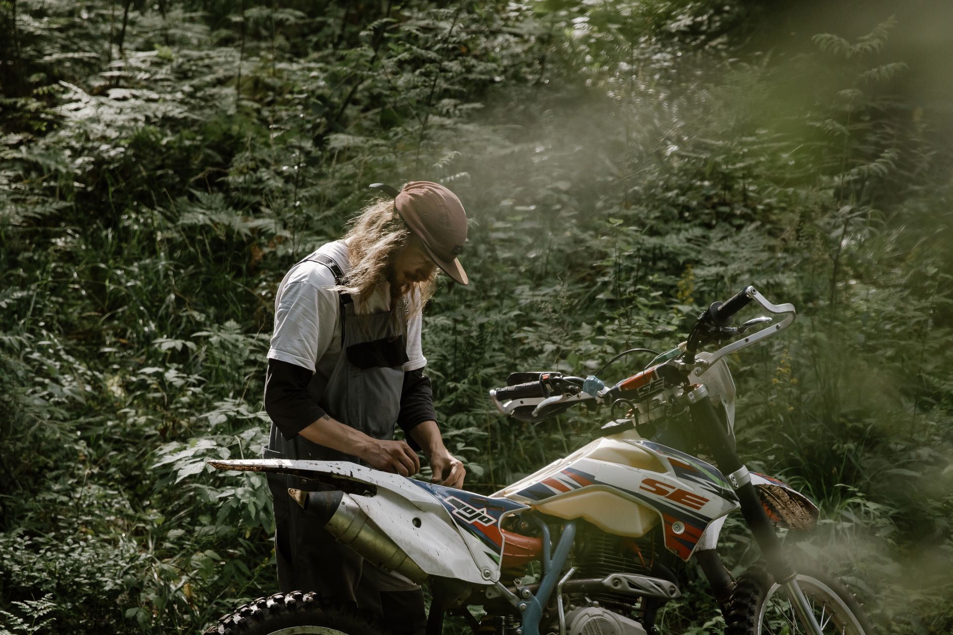A man with long hair repairs his dirt bike in a lush, outdoor forest setting during the day.