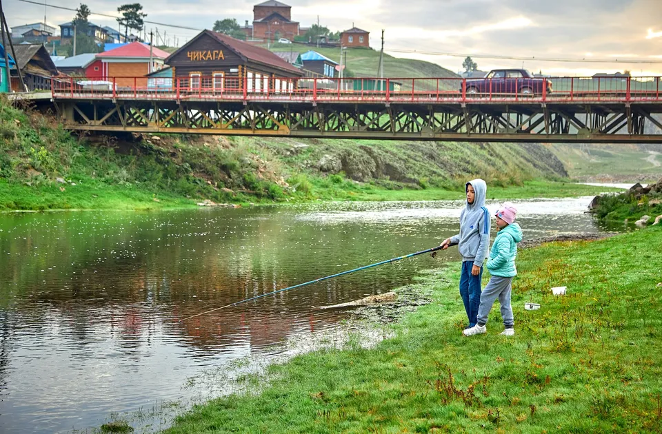 Two children fishing by a river under a bridge in a serene natural setting.